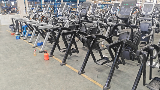 Row of commercial elliptical machines in a workshop or warehouse setting undergoing maintenance or refurbishment, with cleaning bottles and tools placed nearby.