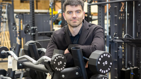 Tim French in a black blazer standing in a gym equipment showroom, leaning on a rack of French Fitness dumbbells and curl bars