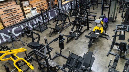 Large warehouse filled with commercial gym equipment including leg press machines, squat racks, and strength training stations, with boxed French Fitness gear stacked on shelves in the background.