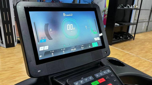 Close-up of a treadmill touchscreen console displaying workout data and controls, with a large red stop button and exercise equipment visible in the background.