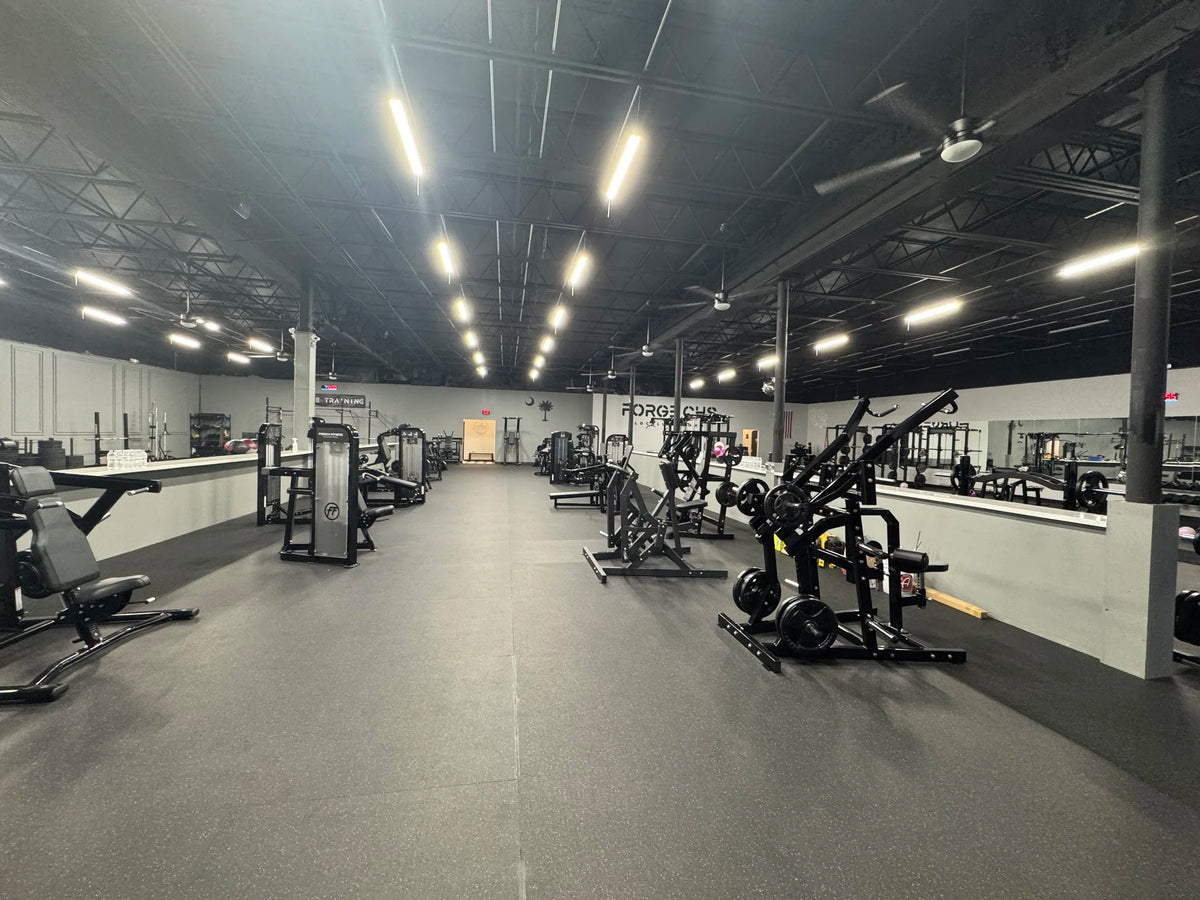 Wide open gym floor at Forge CHS with squat racks, plate-loaded machines and benches arranged along a mirrored wall under overhead strip lighting