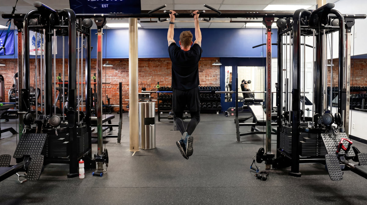 Gym member performing pull-ups on a bar between two multi-station cable machines, with dumbbell racks and benches along a brick wall behind